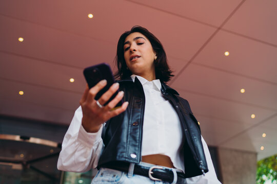 Spanish female student checking transport schedules smartphone application. Hispanic woman text messaging on mobile phone screen device near modern architectural pavilion building corridor.