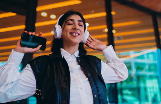Hispanic woman closing eyes enjoying music stereo sound track app. Joyful spanish female digital nomad holding smartphone tool while dancing with wireless headphones in terminal lobby area.