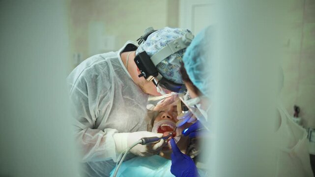  A dentists drilling a hole for a dental implant in the jaw of a young adult female patient with a dental drill, while draining excess saliva. Footage of a real-life surgical procedure