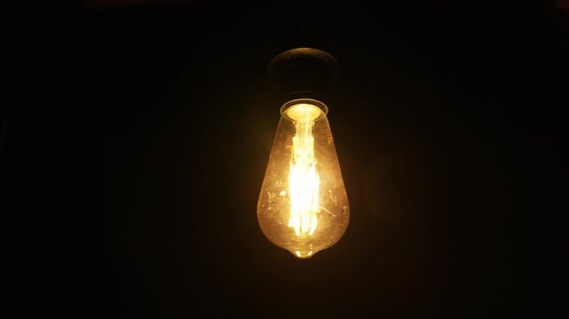 A retro filament light bulb hangs suspended and glows intensely. Warm yellow light radiates from the glass bulb against a completely dark background.