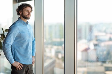 Fototapeta premium Focused Man In Mid-30s, Caucasian, Contemplating While Looking Out Large Window In Contemporary Office Space, Reflecting On Challenges And Opportunities. Business Concept.