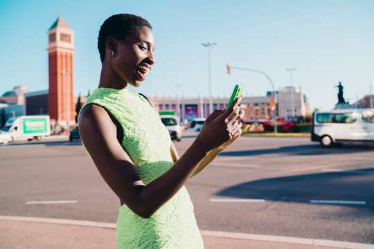 Side view of laughing dark skinned woman using smartphone for online chatting on sunlit city background. Happy African American youth reading funny news or browsing media application outdoors.