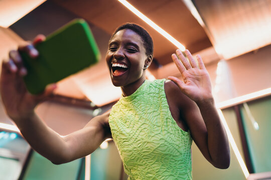 Joyful African American woman waving hand to mobile camera while capturing selfie video chat. Happy dark skinned female blogger using wireless connection under interior neon lights.
