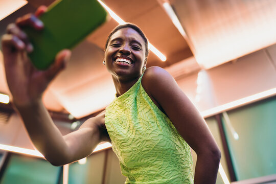 Low angle portrait of happy African American woman browsing internet network on green smartphone. Cheerful dark skinned female youth looking at camera with radiant smile inside terminal.