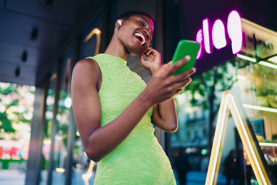 Overjoyed black woman holding green smartphone and laughing out loud near purple neon sign. Happy African American female model receiving great news or winning opportunity online.