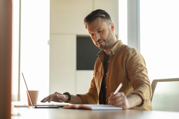 Fototapeta premium Focused Businessman In Casual Attire Writing Notes While Working On Laptop At Modern Office Setting, Displaying Concentration And Professionalism. Engaged In Productive Business Concept.