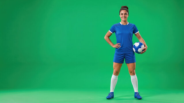 A smiling female soccer player stands against a green screen. She wears a blue uniform, white socks, and cleats, holding a soccer ball.