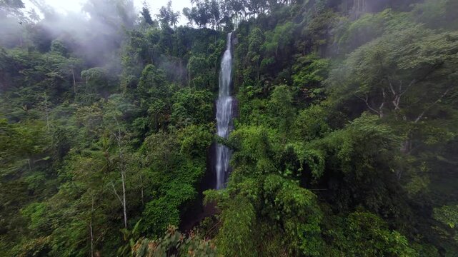 Cinematic FPV drone flight toward a tall tropical waterfall surrounded by lush rainforest in Java, Indonesia. Scenic aerial view of wild nature, jungle landscape and flowing water.
