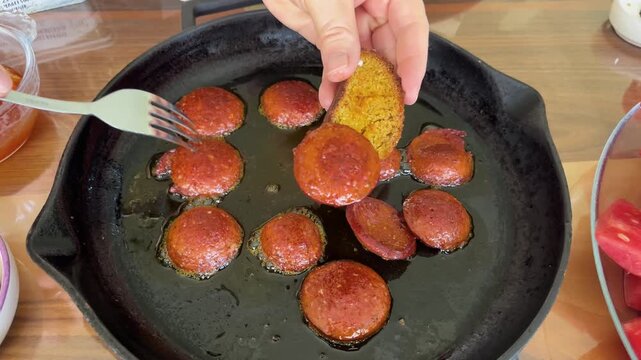 Delicious sucuk sizzling in a cast-iron skillet and a hot slice of sucuk placed on top of crispy bread with the help of a fork. A delicious traditional breakfast moment.
