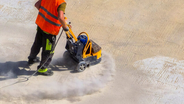 Worker wears bright orange vest. He pushes yellow concrete saw. Dust sprays from the cut. He concentrates on the task. Concrete surface shows work progress. Ideal for construction projects