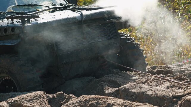 Extreme close up of muddy 4x4 vehicle stuck in deep sand. Smoke billows from overheated engine during intense off road competition. Winch cable pulls powerful truck through difficult obstacle