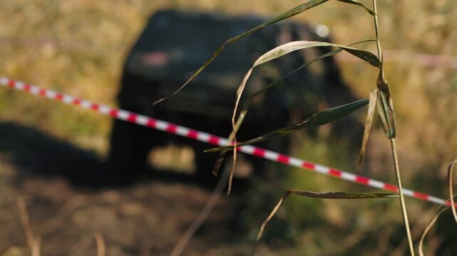 Close up focus on foreground grass with blurred off-road truck at extreme competition. Red and white barrier tape separates spectator view. Warm natural light creates gritty, adventurous mood