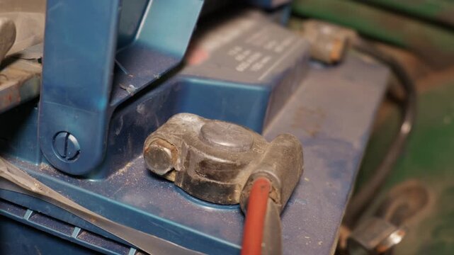 Car battery terminal is connected to a cable clamp inside a vehicle engine compartment. A metal wing nut secures the battery mount while the blue plastic casing shows signs of use.