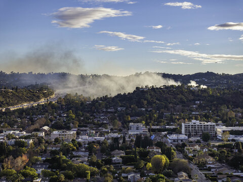 Aerial view of the San Fernando fire burning in the hills above residential neighborhoods with smoke rising into the blue sky over Encino Estates, California, United States.