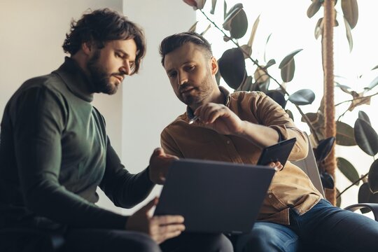 Focused Men Engaged In Discussion Over Laptop Screen In Office Setting, Working Together On Project. Professional Environment Reflecting Teamwork And Collaboration In Business Concept.