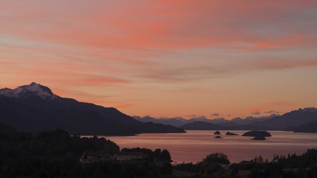 Panoramic wide shot of Lake Nahuel Huapi and hotel nestled among forested hills under a dramatic orange and pink sunset sky in Bariloche, Argentina.