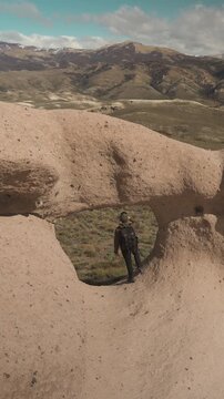 Male hiker with a backpack standing and looking through a natural stone arch hole, with a dry valley and mountain range under a cloudy sky in the background.