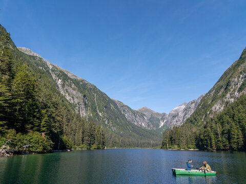 Aerial view of a calm lake surrounded by dense pine forests and towering mountains with two people rowing in a small green boat in Sitka, Alaska, United States.