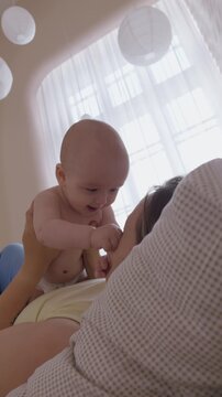 mother lifting baby above chest on bed. morning exercise and play mom with infant with bright window light. cheerful movement and eye contact. happy domestic maternal routine. cozy bonding