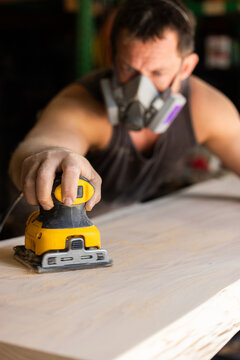 Man wears protective mask while sanding wood with an orbital sander. Focused expression shows care and precision in work.