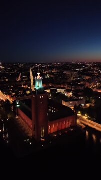 High-angle perspective of Stockholm City Hall tower illuminated at dusk. Professional night drone photography showcasing Scandinavian architecture and the M&auml;laren lake horizon.