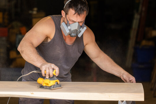 Man wears protective mask while sanding wood with an orbital sander. Focused expression shows care and precision in work.