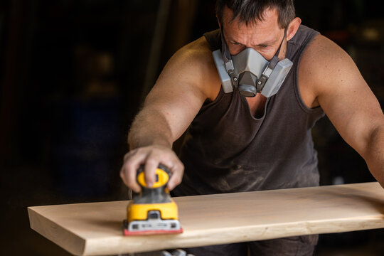 Man wears protective mask while sanding wood with an orbital sander. Focused expression shows care and precision in work.