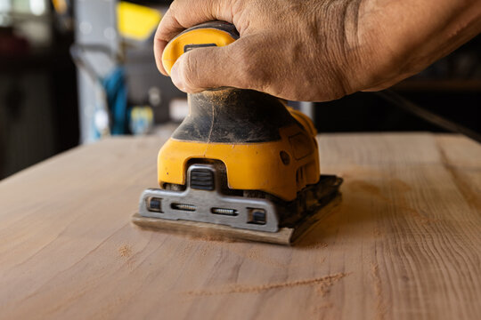Hand guides power orbital sander over wooden surface. Workbench hints at craftsmanship or DIY project