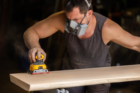 Man wears protective mask while sanding wood with an orbital sander. Focused expression shows care and precision in work.