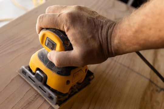 Hand guides power orbital sander over wooden surface. Workbench hints at craftsmanship or DIY project