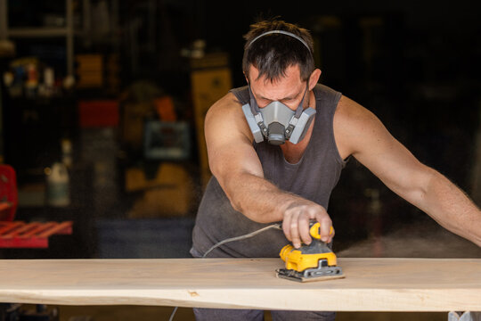 Man wears protective mask while sanding wood with an orbital sander. Focused expression shows care and precision in work.