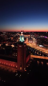 High-angle perspective of Stockholm City Hall tower illuminated at dusk. Professional night drone photography showcasing Scandinavian architecture and the M&auml;laren lake horizon.