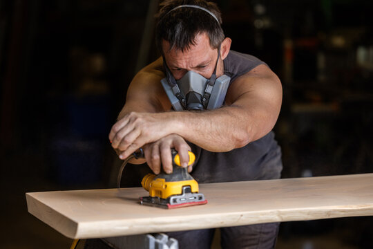 Man wears protective mask while sanding wood with an orbital sander. Focused expression shows care and precision in work.