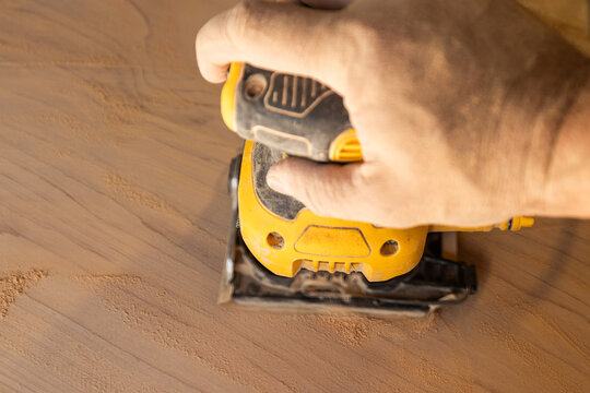 Hand guides power orbital sander over wooden surface. Workbench hints at craftsmanship or DIY project