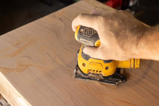 Hand guides power orbital sander over wooden surface. Workbench hints at craftsmanship or DIY project