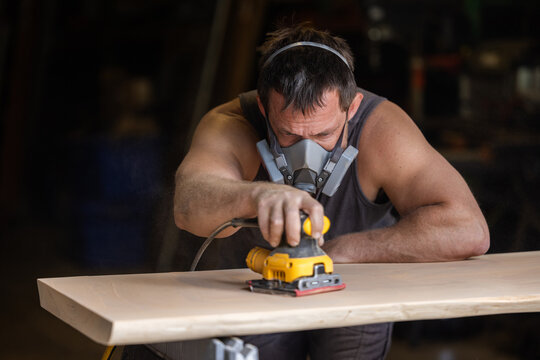 Man wears protective mask while sanding wood with an orbital sander. Focused expression shows care and precision in work.