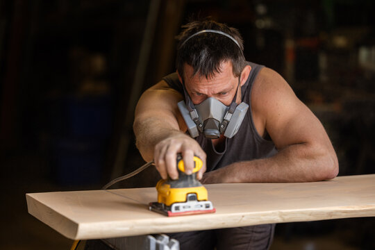 Man wears protective mask while sanding wood with an orbital sander. Focused expression shows care and precision in work.