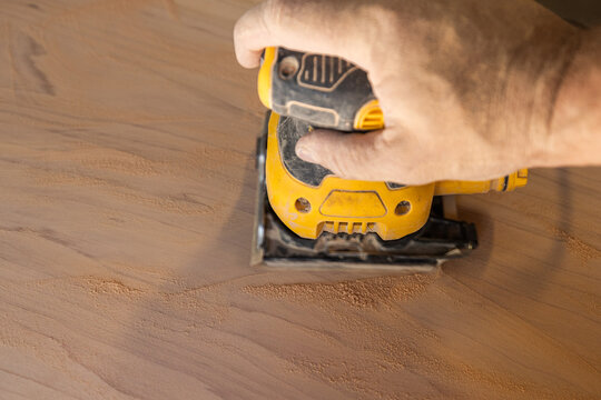 Hand guides power orbital sander over wooden surface. Workbench hints at craftsmanship or DIY project