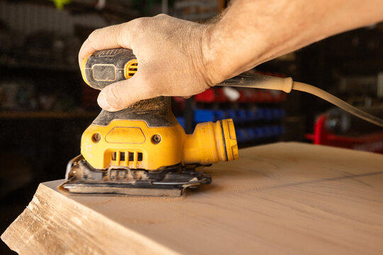 Hand guides power orbital sander over wooden surface. Workbench hints at craftsmanship or DIY project