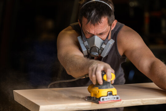 Man wears protective mask while sanding wood with an orbital sander. Focused expression shows care and precision in work.