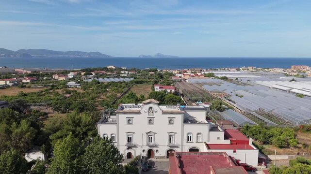 Aerial drone view of historic white villa surrounded by greenhouses and trees with tyrrhenian sea and islands in background, Torre del Greco, Naples, Italy
