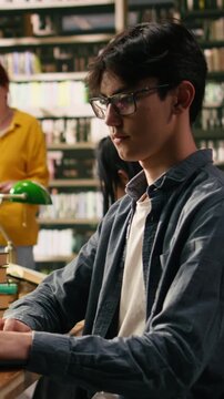 Vertical frame view: Asian student working on laptop in quiet library with tall bookshelves and blurred classmates, warm lighting for academic study environment, Independent study session.