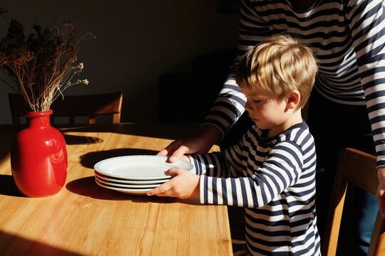 Boy helps adult set table. Child places plates on wooden dining table. Kid learns household chores, domestic routine. Family meal preparation indoors. Casual scene, everyday life.