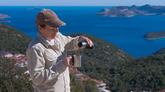Hiker pouring hot coffee from thermos on mountain top. Female traveler pouring a hot drink from a thermos into a mug, enjoying the scenic view of the sea and islands from a mountain peak during a hike