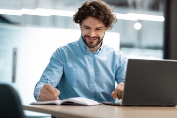 Obraz premium Smiling Caucasian Man Focused On Writing Notes While Working On Laptop At Modern Office Desk, Engaged In Productive Activity, Creating Ideas For Project, Business Concept.