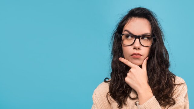 Contemplative Young Woman With Glasses, Deep In Thought While Pondering Ideas In A Studio Setting Against A Blue Background. Reflecting On Business Decisions And Strategies.