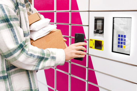 Person holding packages and using a smartphone at an automated parcel locker terminal. Convenient contactless pickup and delivery service for online ecommerce shopping logistics.