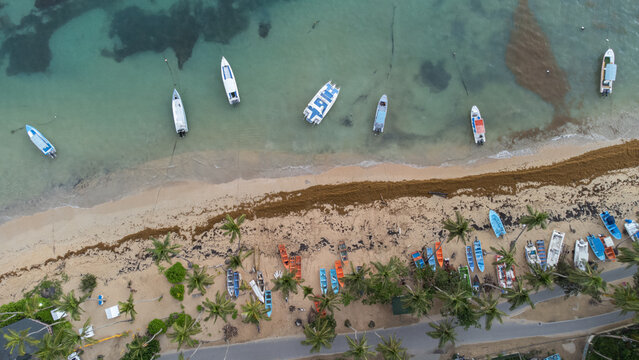 Aerial view of small boats anchored in turquoise waters along a sandy beach lined with palm trees and a coastal road in the Dominican Republic.