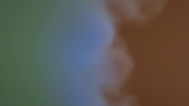 Abstract background featuring a smooth blurry gradient transitioning from muted olive green through soft blue to warm brown and tan earth tones in a slow motion video sequence.