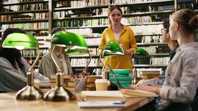 Mentor at group study speaking with peers seated at library table with green lamps, collaborative learning and academic guidance.